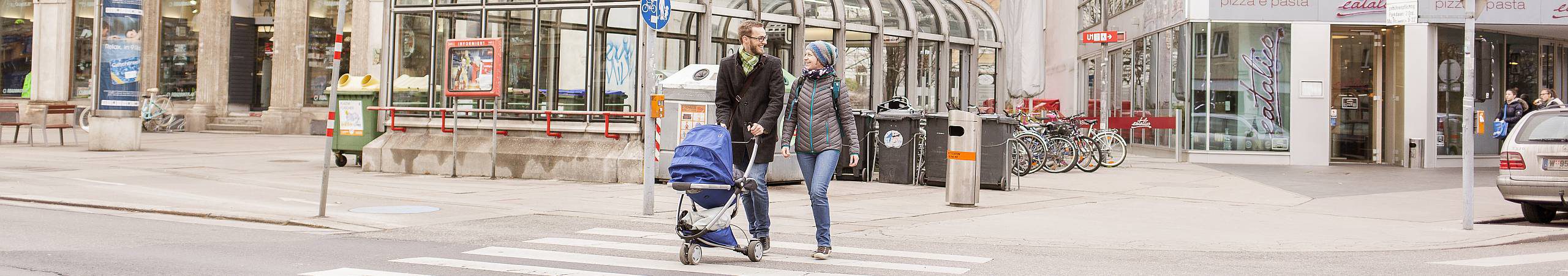 Ein lachendes Paar überquert mit Kinderwagen eine Kreuzung. Foto von Stephan Doleschal