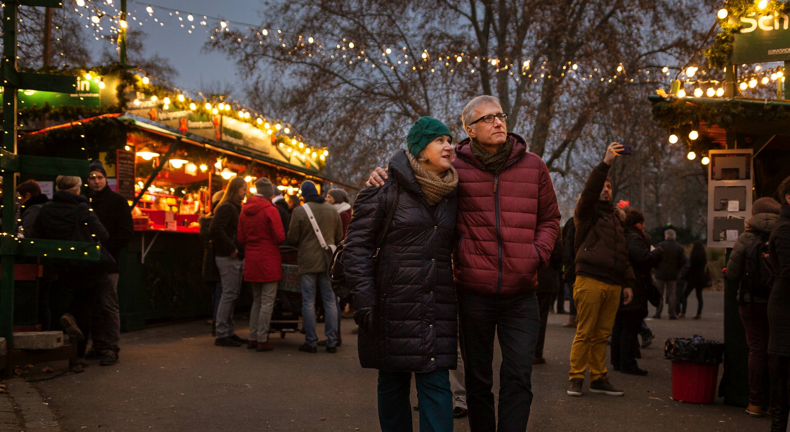 Ein Pärchen mittleren Alters in Winterkleidung flaniert Arm in Arm über einen Weihnachtsmarkt. Im Hintergrund sieht man bunt beleuchtete Marktstände. Foto: Stephan Doleschal