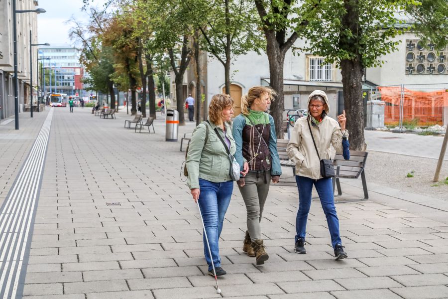 Drei Frauen gehen die Pius-Parsch-Promenade in Floridsdorf entlang. Eine von ihnen hat einen weißen Stock. 