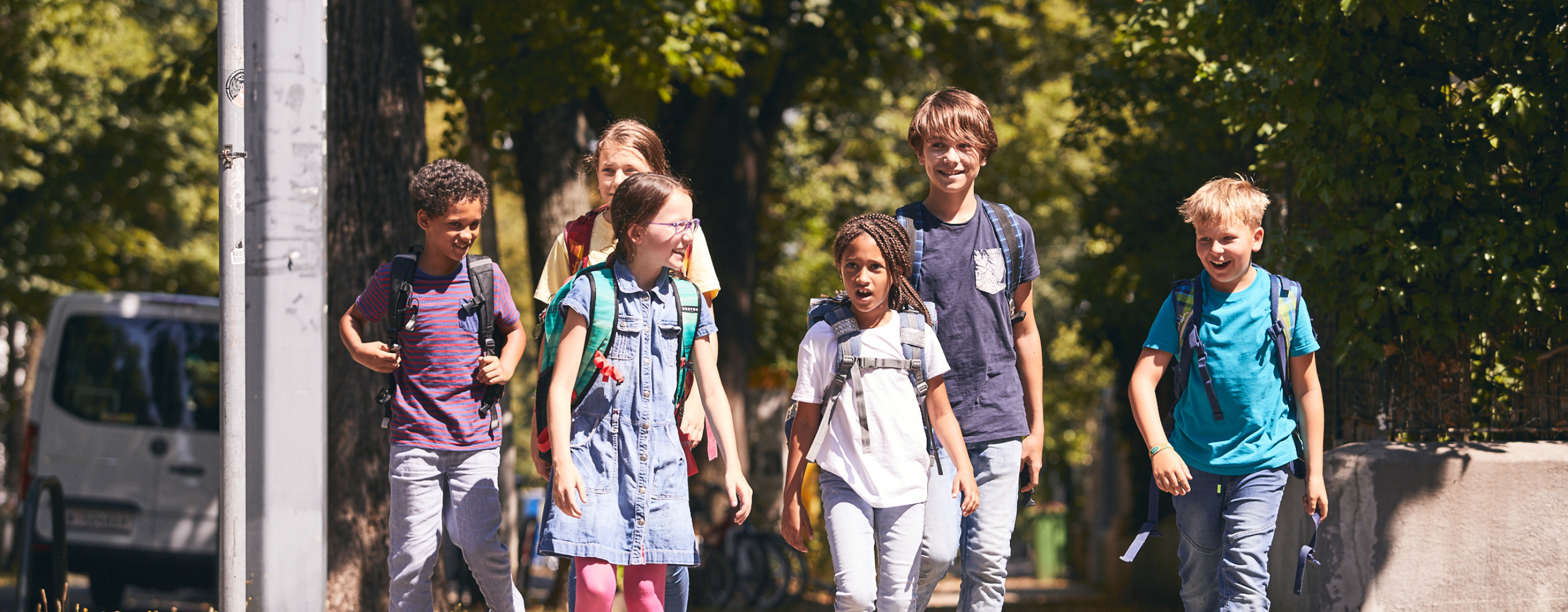 5 Kinder auf ihrem Schulweg, den sie zu Fuß zurücklegen. Sie warten an einer ampelgeregelten Kreuzung.