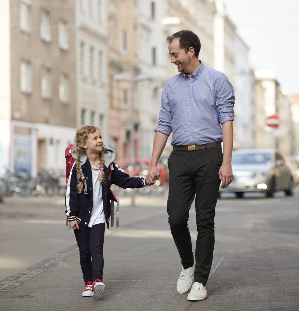 Ein Mann begleitet ein kleines Mädchen zur Schule. Er hält sie an der Hand, sie hat einen Schulrucksack auf dem Rücken.