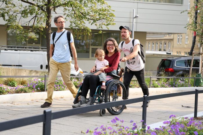 Eine Frau im Rollstuhl mit einem blonden Kind auf dem Schoß. Der Rollstuhl wird von einem jungen Mann mit Mütze geschoben. Ein Mann mit gelber Haus geht neben dem Rollstuhl. Im Vordergrund blühen lila Blumen.