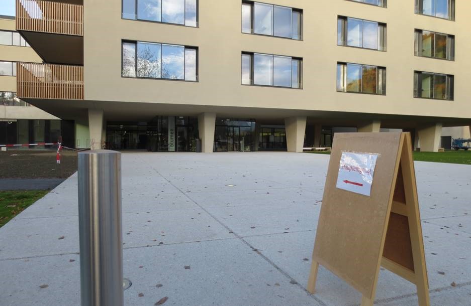 Der Platz vor einem Hochhaus-Gebäude. Auf dem Platz steht ein A-Ständer mit einem Hinweisschil darauf angebracht. Auf dem Hinweisschild ist ein Pfeil und das Wort Haupteingang zu lesen.