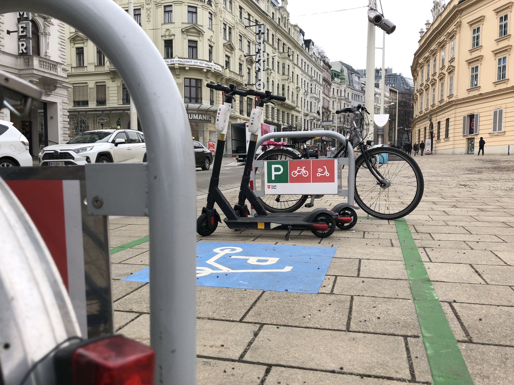 Am Beginn der Mariahilfer Straße ist ein Abstellplatz für e-Scooter markiert. Er ist durch zwei Radbügel begrenzt. An den Bügeln parken Fahrrädern, am Abstellplatz zwei e-Scooter.